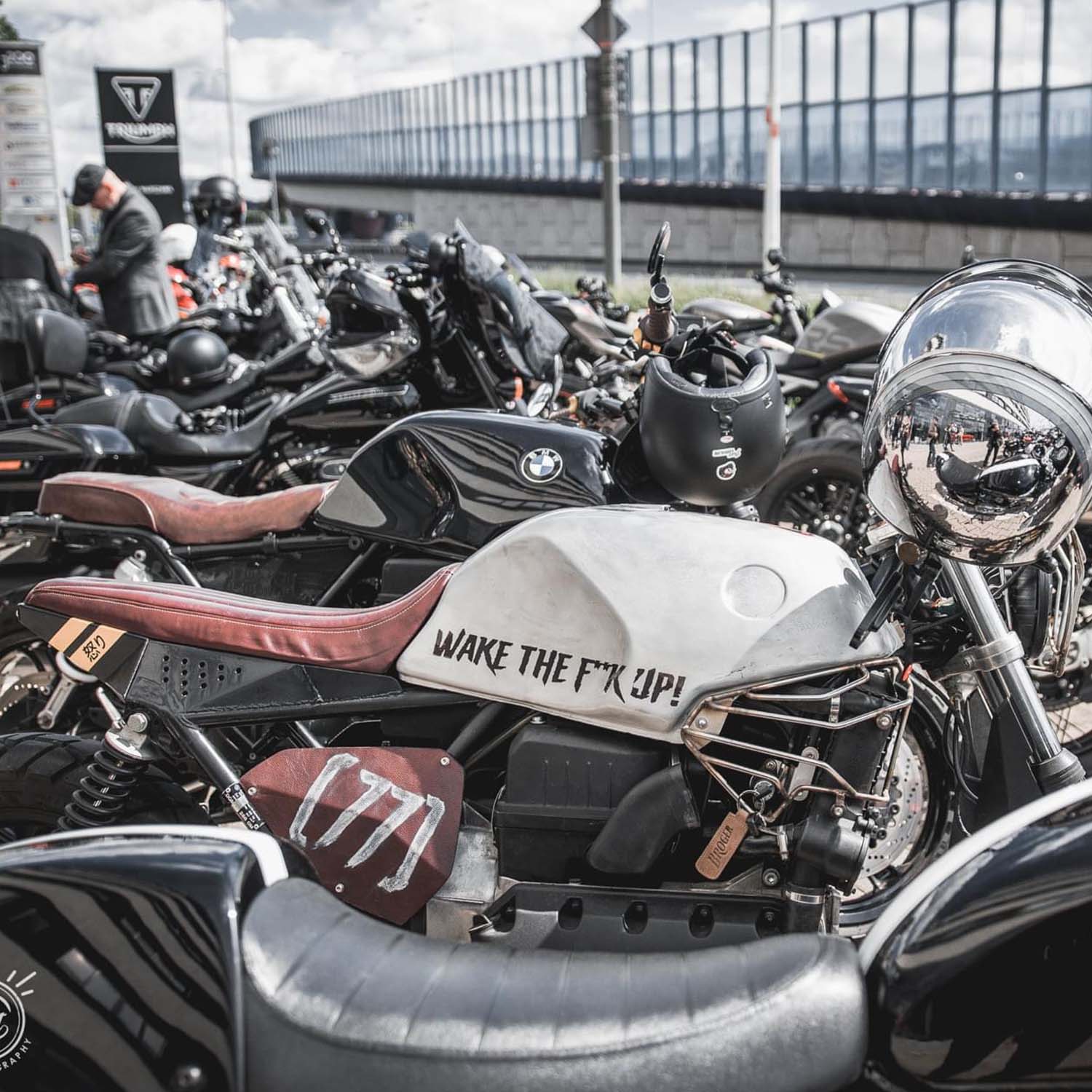 A BMW K-series cafe racer with a brushed silver tank reading WAKE THE F*K UP and a brown leather seat