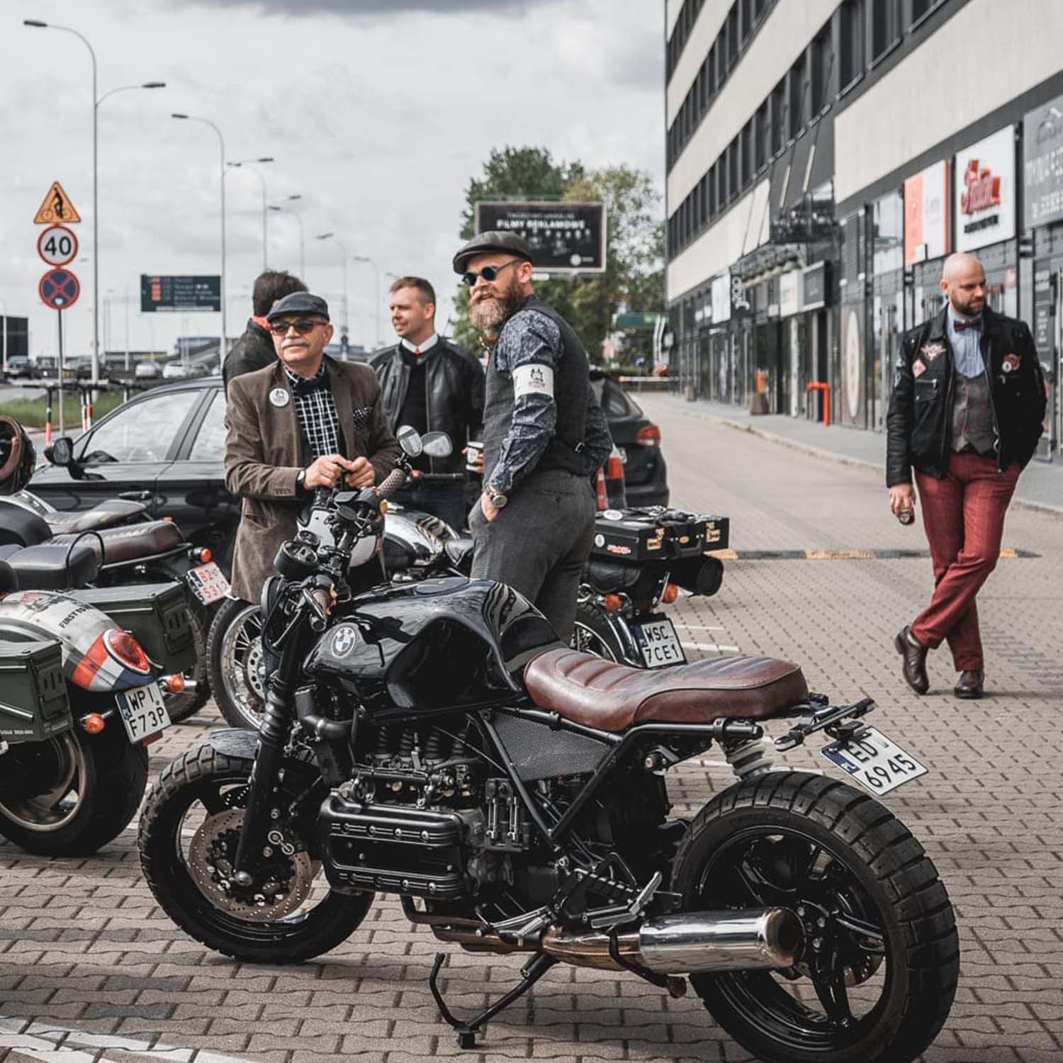 Riders in tweed standing around a BMW K-series brat parked on the street